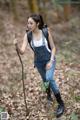 A woman in overalls and boots holding a stick in the woods.